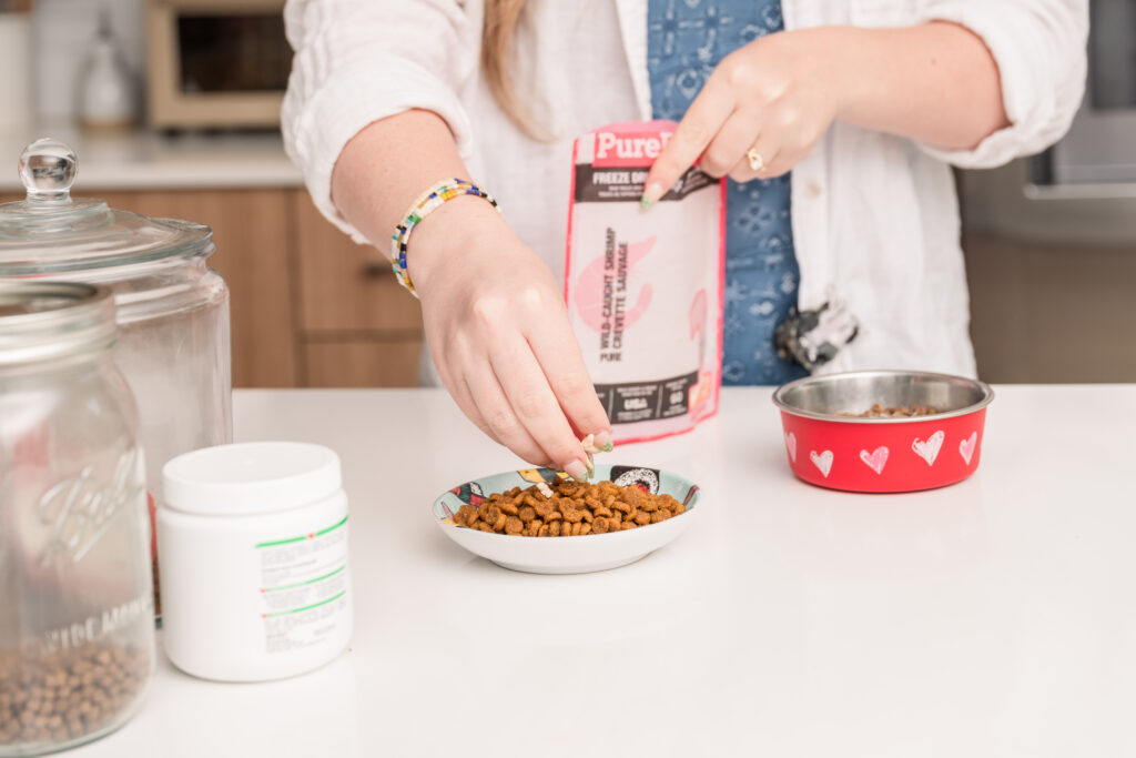 Person preparing pet food by sprinkling freeze-dried shrimp over dry kibble in a decorative dish.
